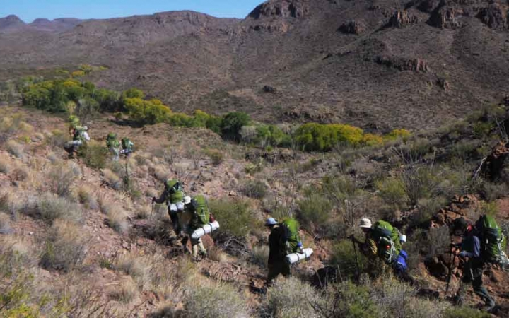 a group of gap year students carrying backpacks hike through a desert landscape on an outward bound semester course